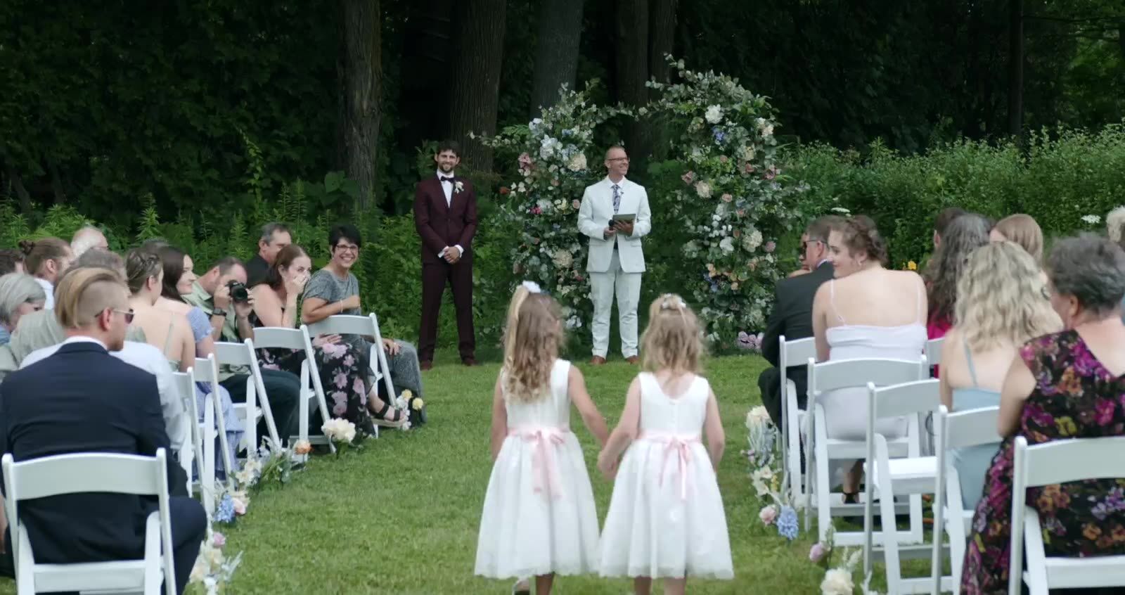 Two flower girls walking down a garden aisle toward the ceremony arch